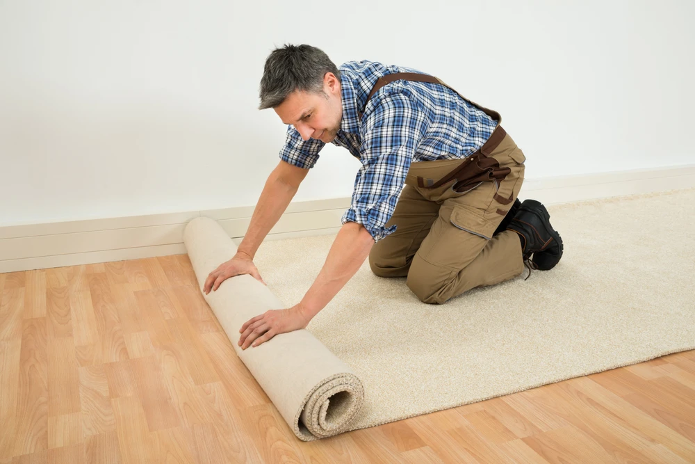Un homme en chemise à carreaux et salopette beige déroule un tapis beige dans une pièce aux murs blancs sur parquet bois clair